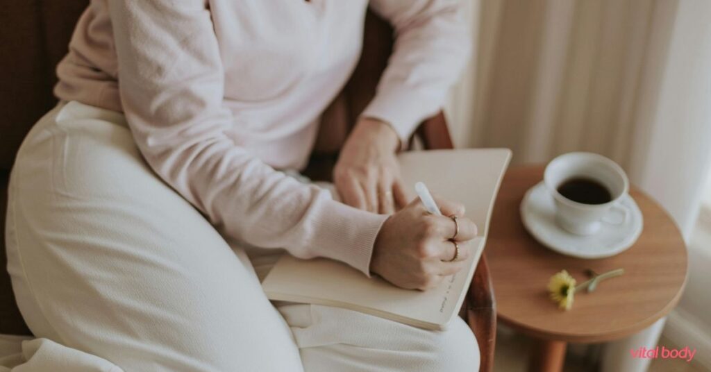 A woman in a cosy chair writing in a food journal with a cup of coffee beside her