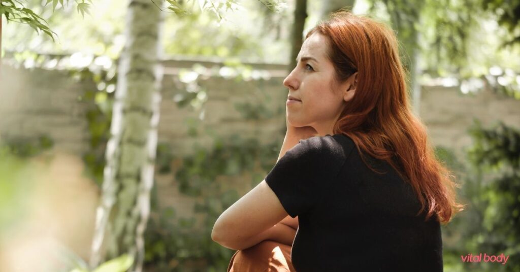 Woman with red hair sitting outdoors among birch trees, looking ahead thoughtfully