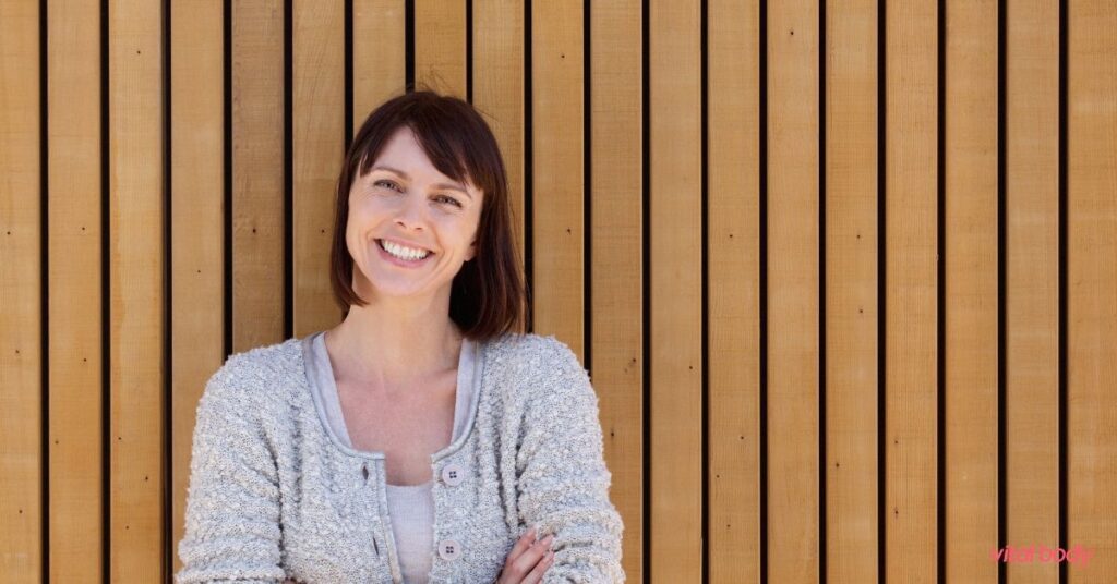 A smiling woman with arms crossed in front of a wooden wall, confident and at home in her body on Ideal Protein Phase 3