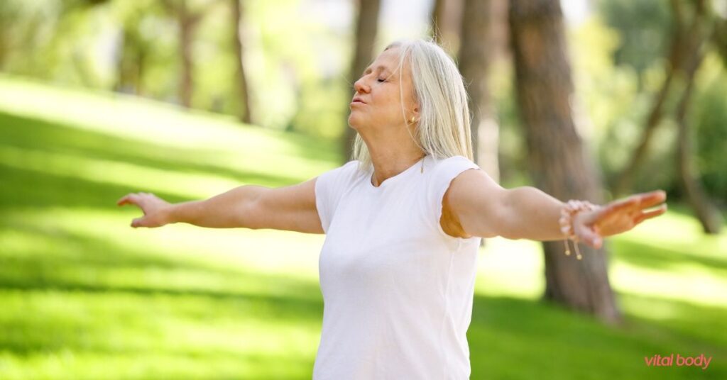 Woman with silver hair standing in a sunny park with her arms open and eyes closed, taking a deep breath