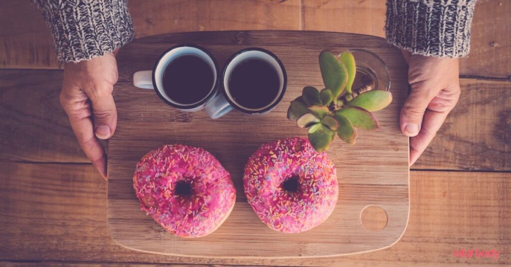 Hands holding a wooden board with two coffees and two pink sprinkled doughnuts