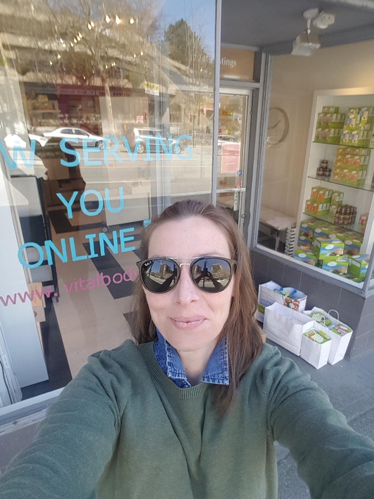 Em, Vital Body coach, outside the clinic window reading Now Serving You Online, with order bags ready for curbside pickup