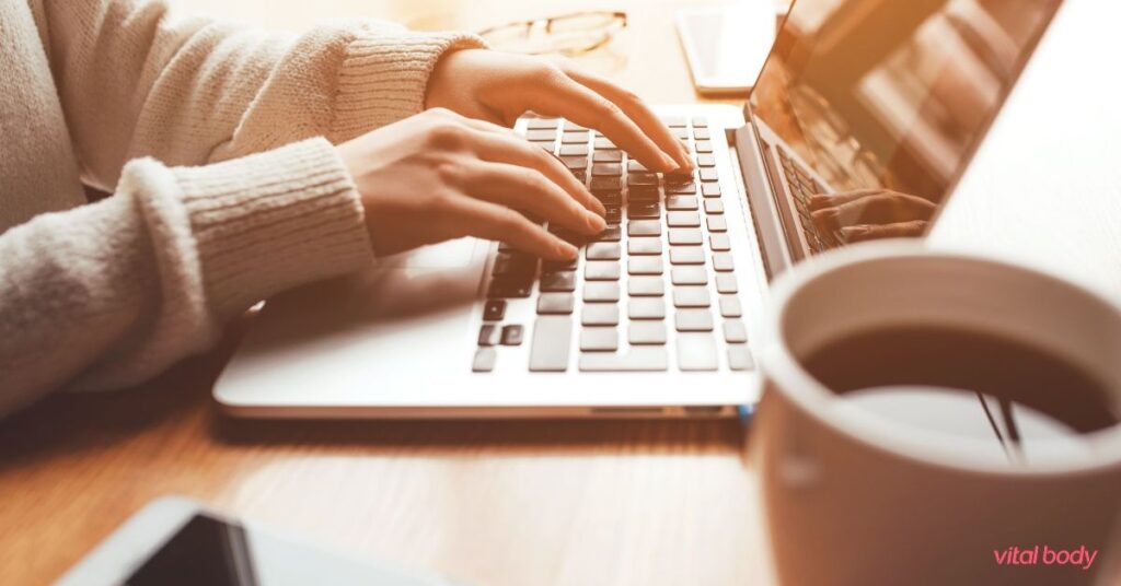 Woman in a cream sweater typing on her laptop at a wooden table, with a cup of coffee beside her